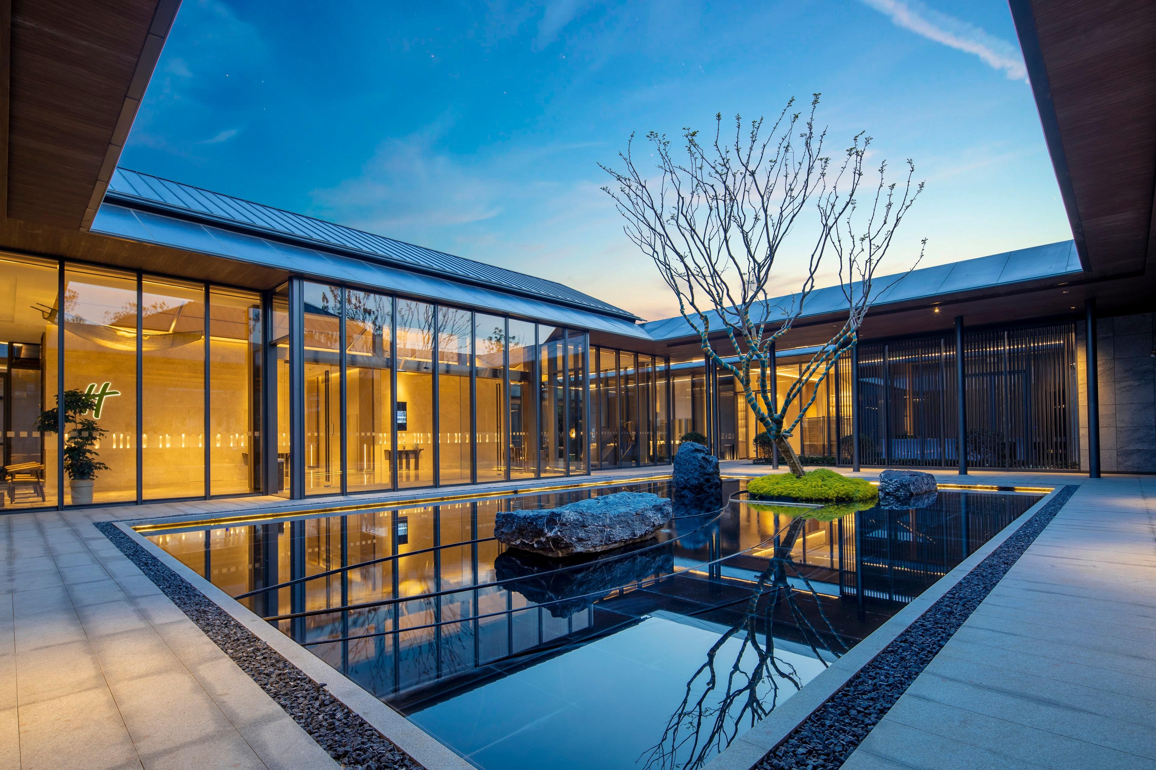 A modern building with a glass facade, a reflective water feature, and a tree in the courtyard.