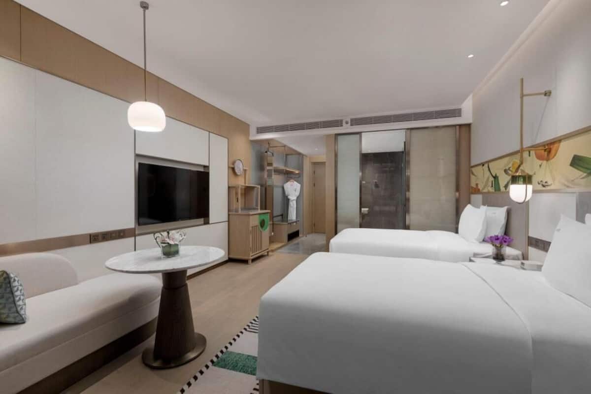 Habitación de hotel con dos camas, un sofá, un televisor, una mesita y un baño visible a través de una puerta de cristal.