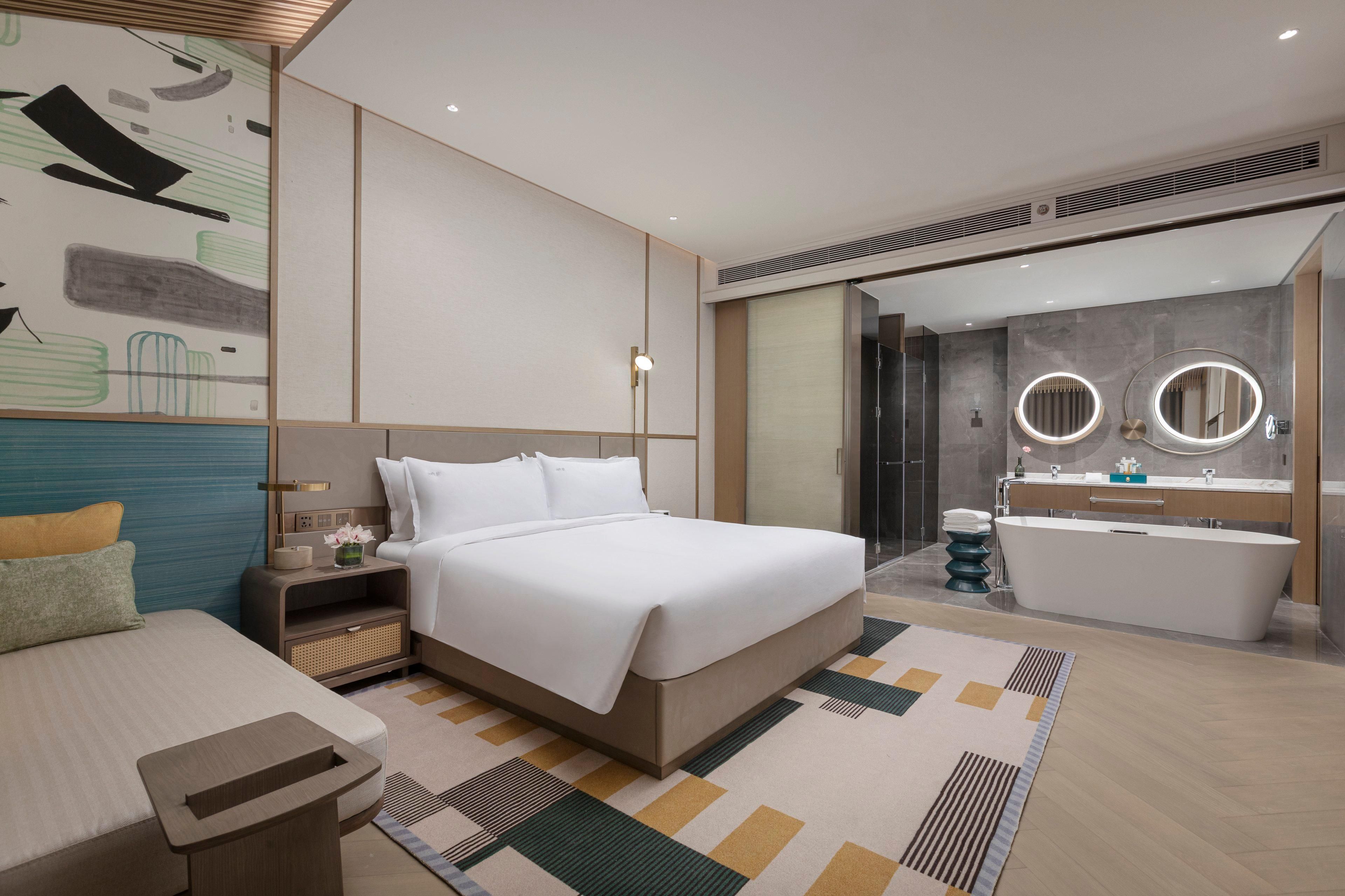 A modern hotel room with a large bed, bedside table, and a bathroom with a bathtub and two round mirrors.