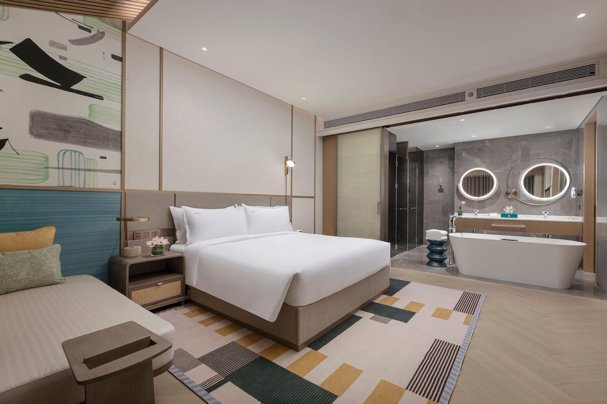 Ein modernes Hotelzimmer mit einem großen Bett, einem Nachttisch und einem Badezimmer mit Badewanne und zwei runden Spiegeln.