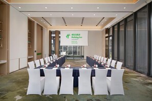 Une salle de conférence avec une longue table préparée pour une réunion, des chaises blanches et une banderole ornée du logo de Holiday Inn.