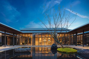 A modern building with a glass facade, a reflective pool, and a tree in the foreground. - Holiday Inn Deqing Xinshi Ancient Town (Huzhou)
