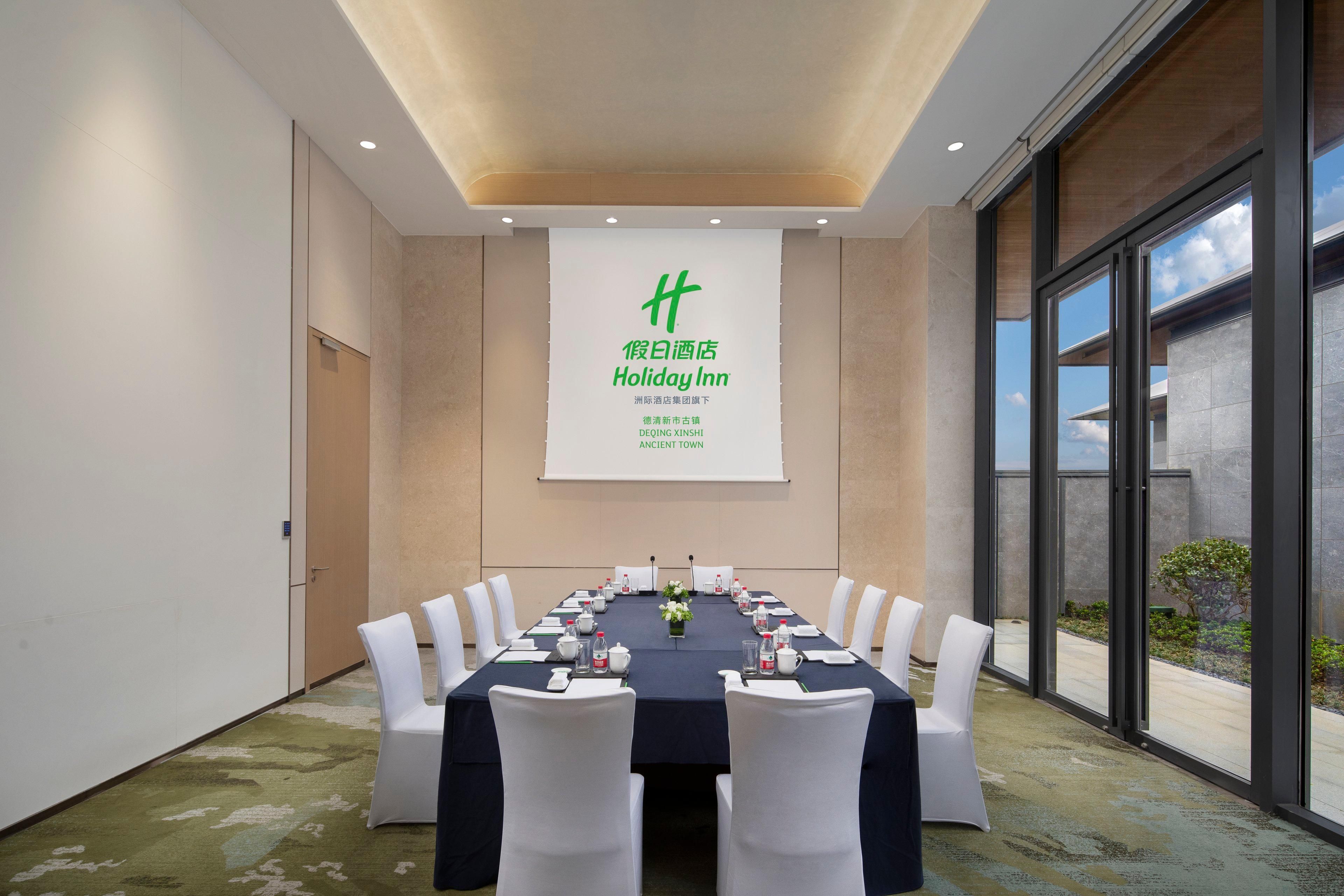 A conference room with a long table set for a meeting, white chairs, and a large screen displaying text.