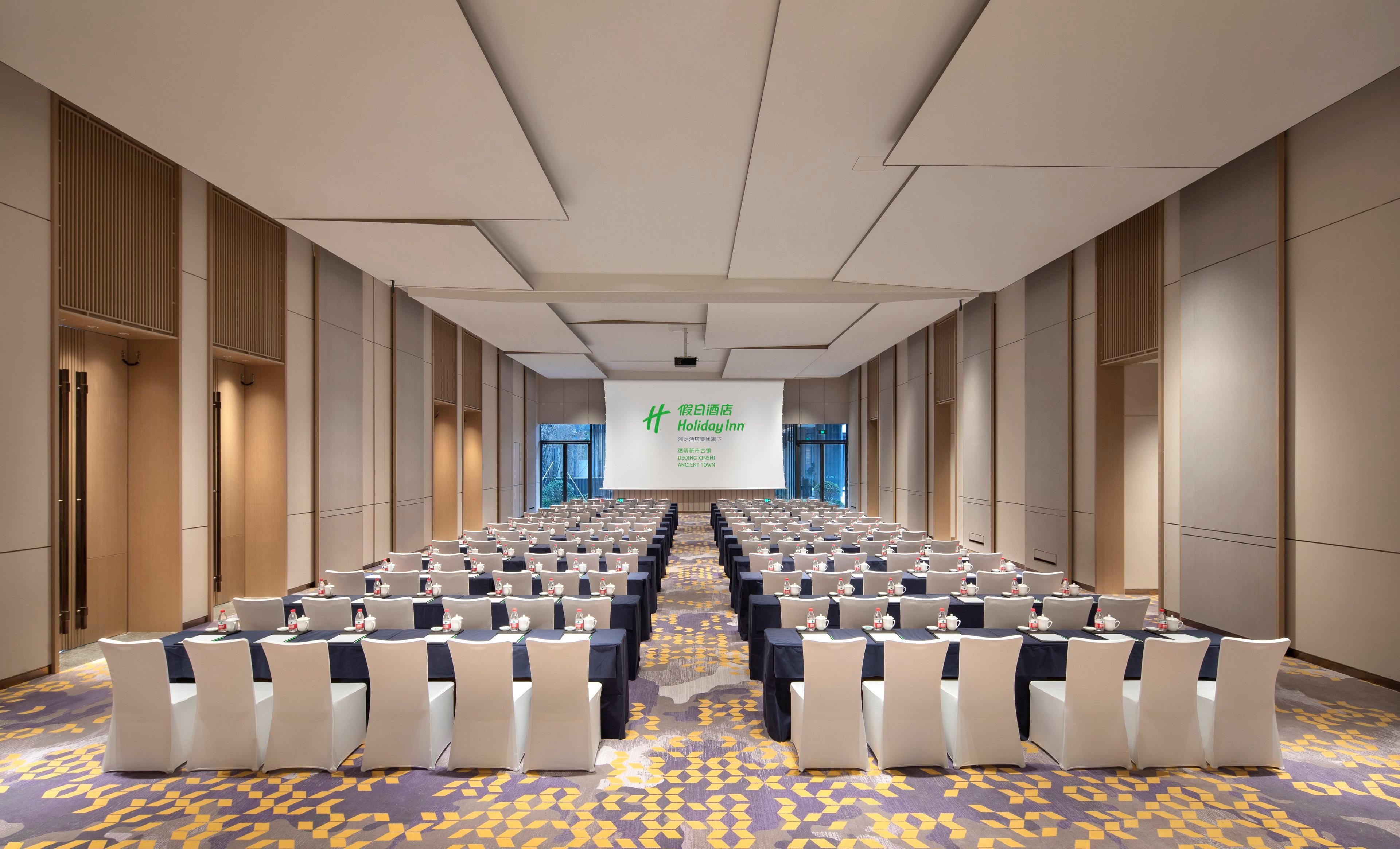 Un salón de conferencias con filas de mesas y sillas, una pantalla grande al frente y una alfombra estampada.
