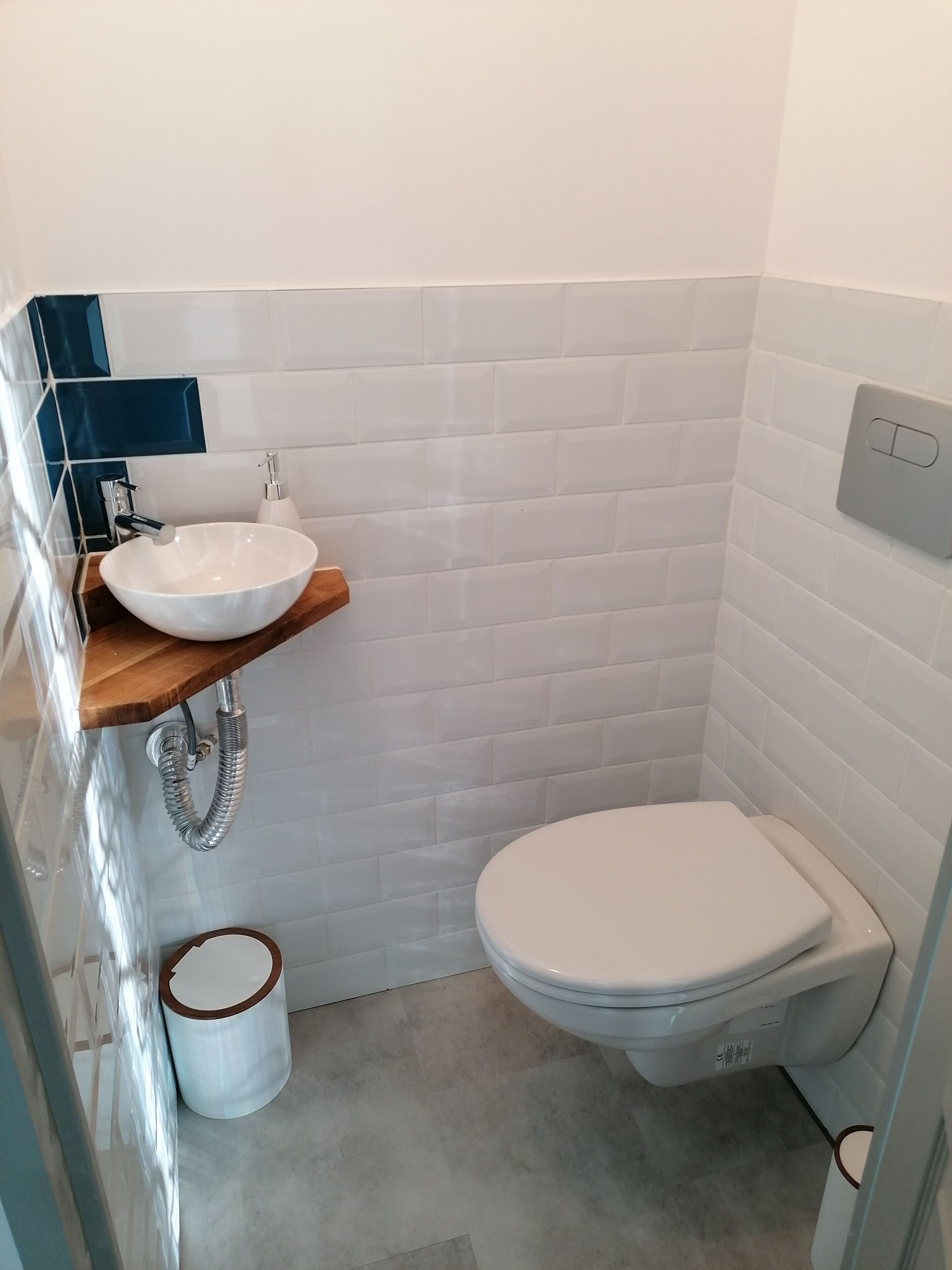 A bathroom with a white toilet, a white sink on a wooden shelf, and a white tiled wall.