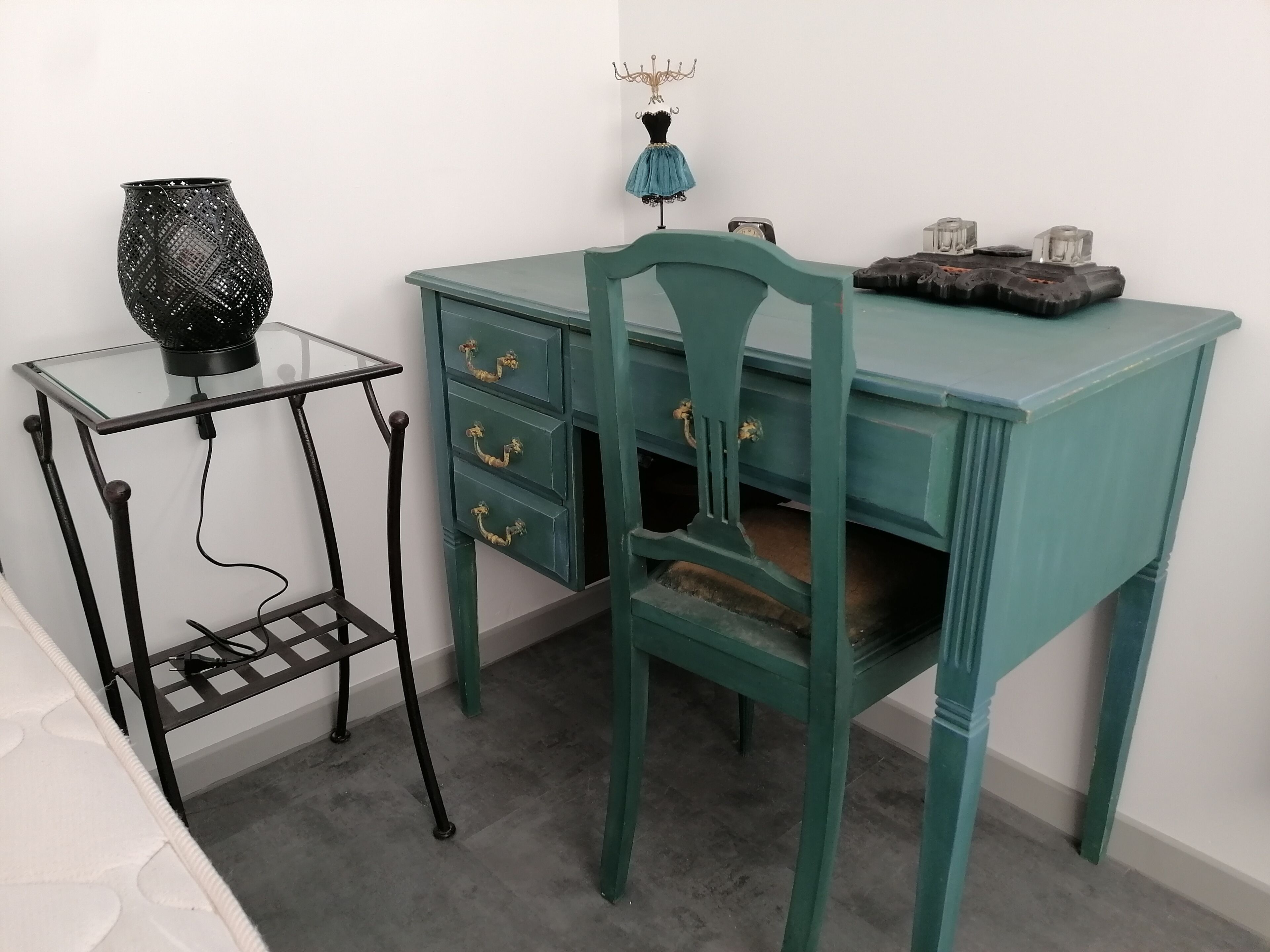 A teal desk with three drawers and a chair, a glass table with a black vase, and a wall-mounted lamp.