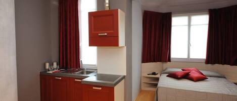 A compact room with a kitchenette, a bed, and a window with curtains.