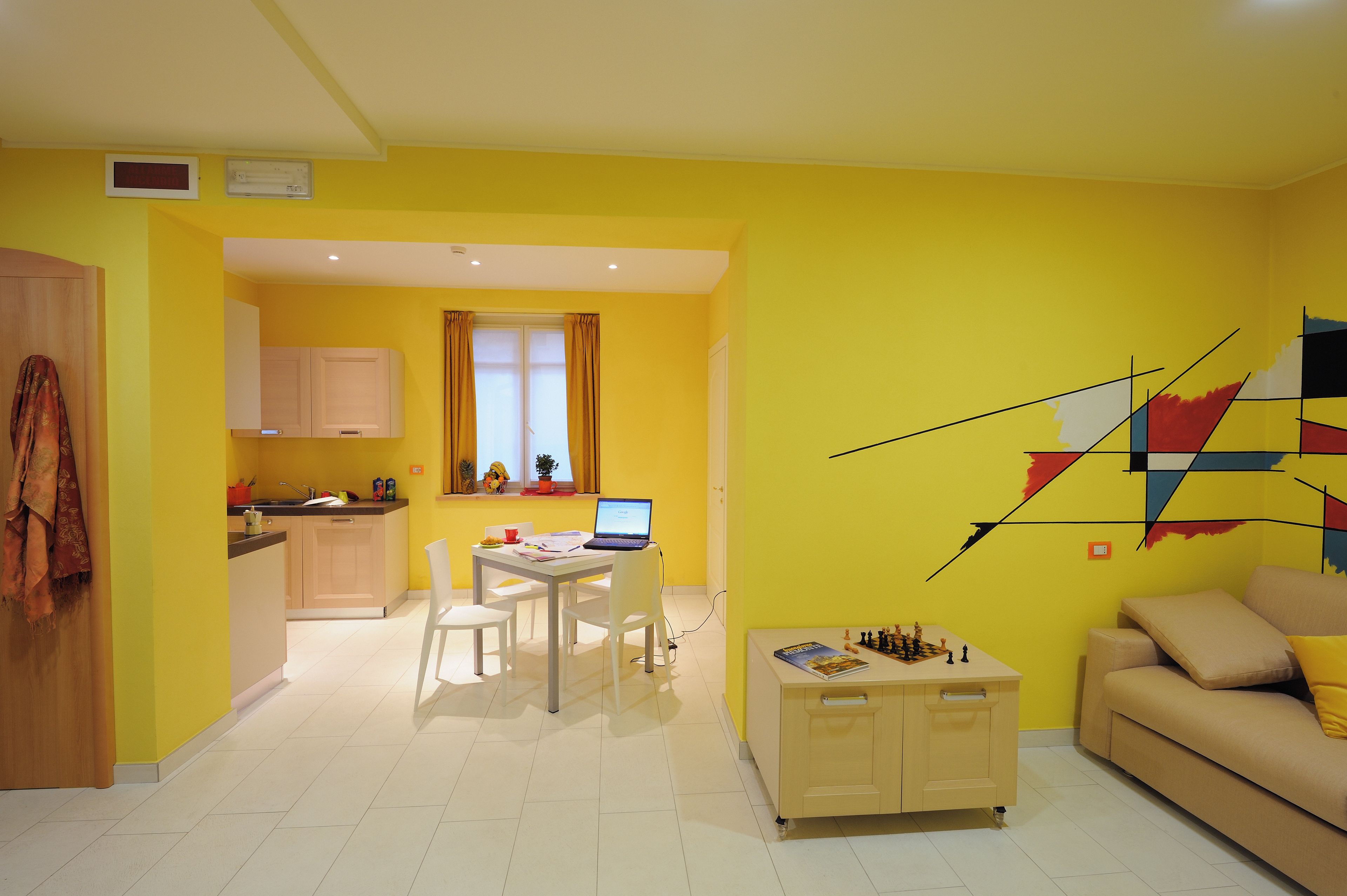 Un soggiorno luminoso e giallo con un divano bianco, un tavolino da caffè e un laptop su un piccolo tavolo. È presente una zona cucina con mobili e una finestra con tende.