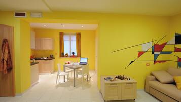 Un soggiorno luminoso e giallo con un divano bianco, un tavolino da caffè e un laptop su un piccolo tavolo. È presente una zona cucina con mobili e una finestra con tende.