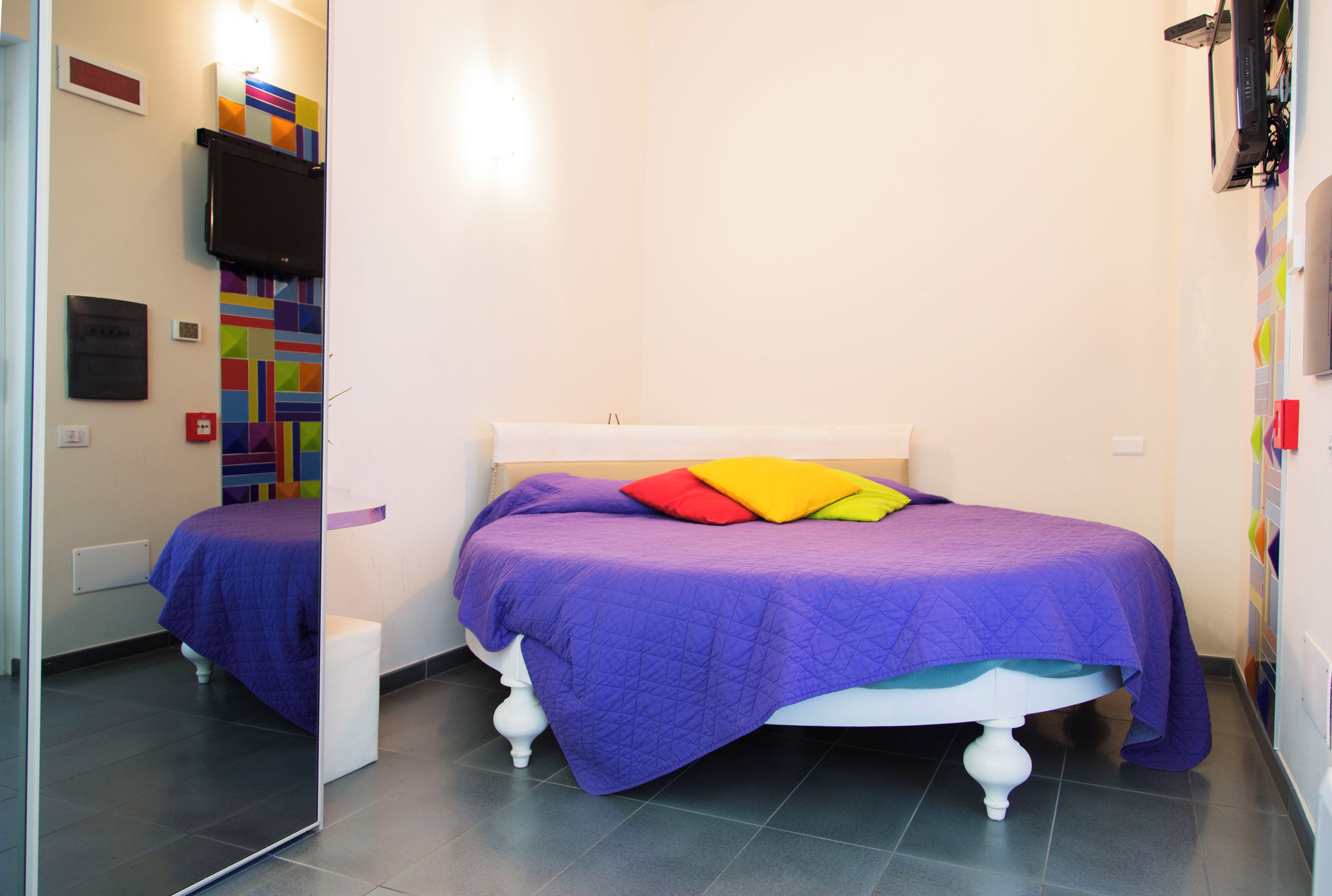 A bedroom with a bed, a small white stool, a wall-mounted TV, and a colorful geometric wall decoration.