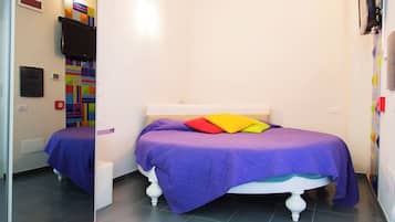 A bedroom with a bed, a small white stool, a wall-mounted TV, and a colorful geometric wall decoration.