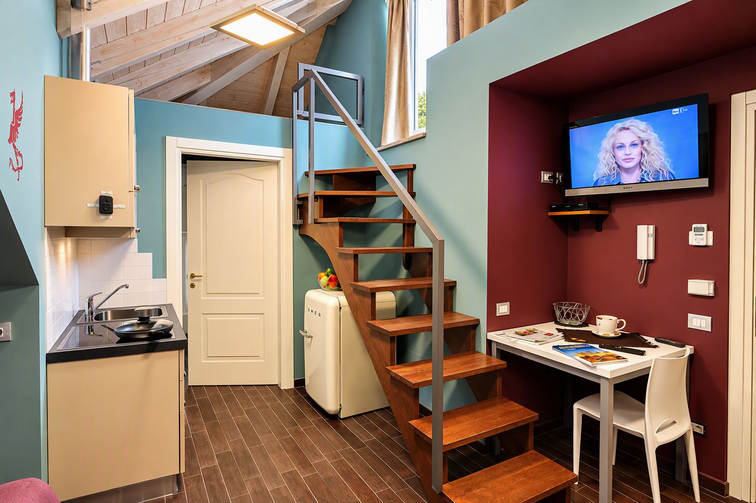 A compact kitchen with a built-in fridge, a wooden staircase, a dining area with a table and chairs, and a television mounted on the wall.
