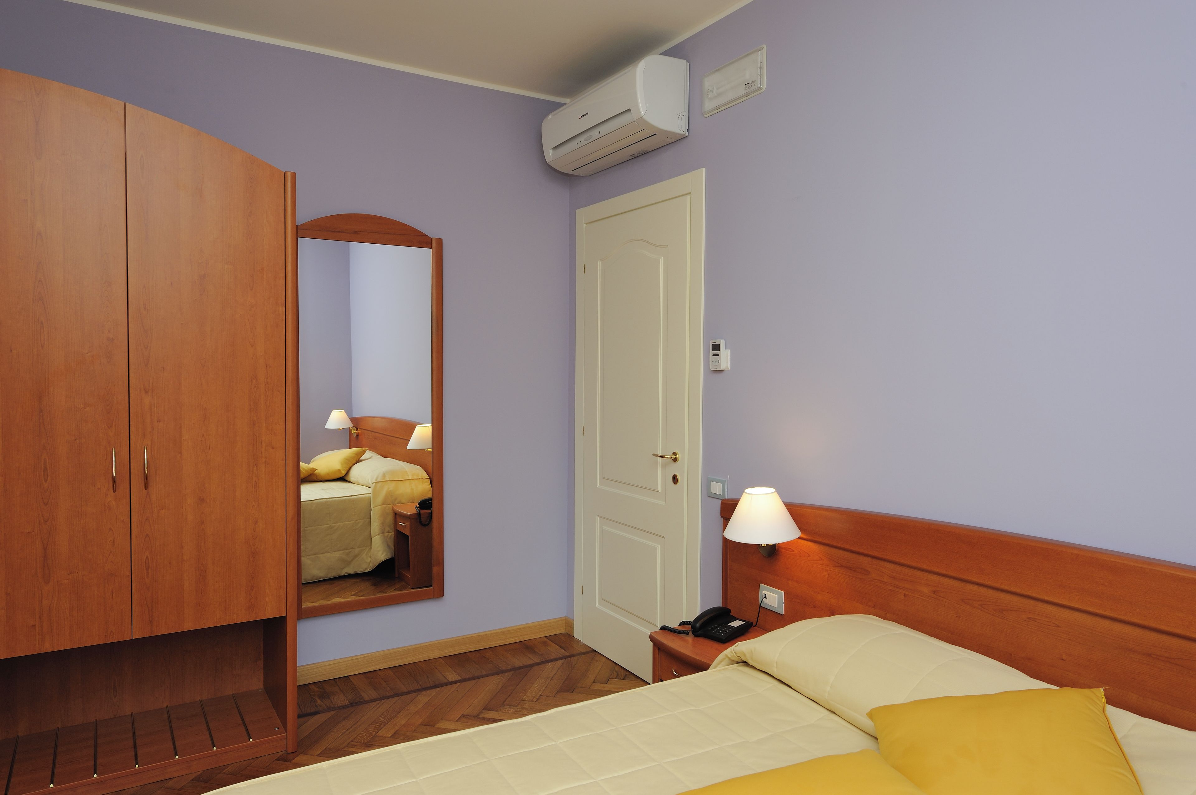 A hotel room with a bed, bedside table, lamp, wardrobe, and an air conditioning unit.