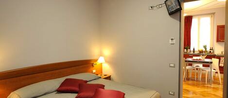 Habitación de hotel con una cama, una mesita de noche con lámpara, cabecero de madera, y vista a la cocina y comedor a través de una puerta abierta.