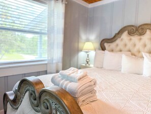 A neatly made bed with a stack of white towels, a wooden headboard, a bedside lamp, and a window with blinds.