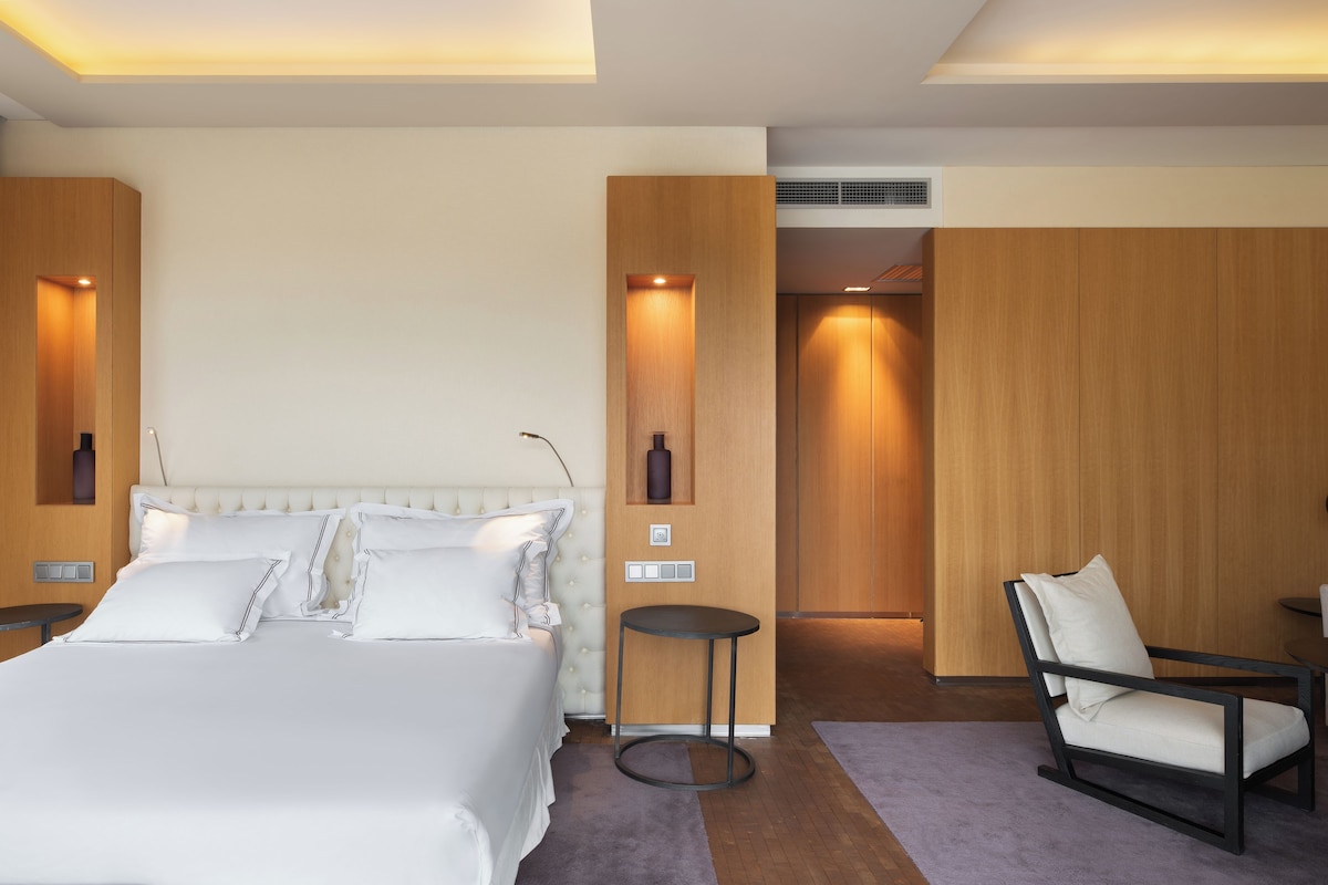 A modern hotel room with a large bed, a small round table, a chair, and a wooden wardrobe.