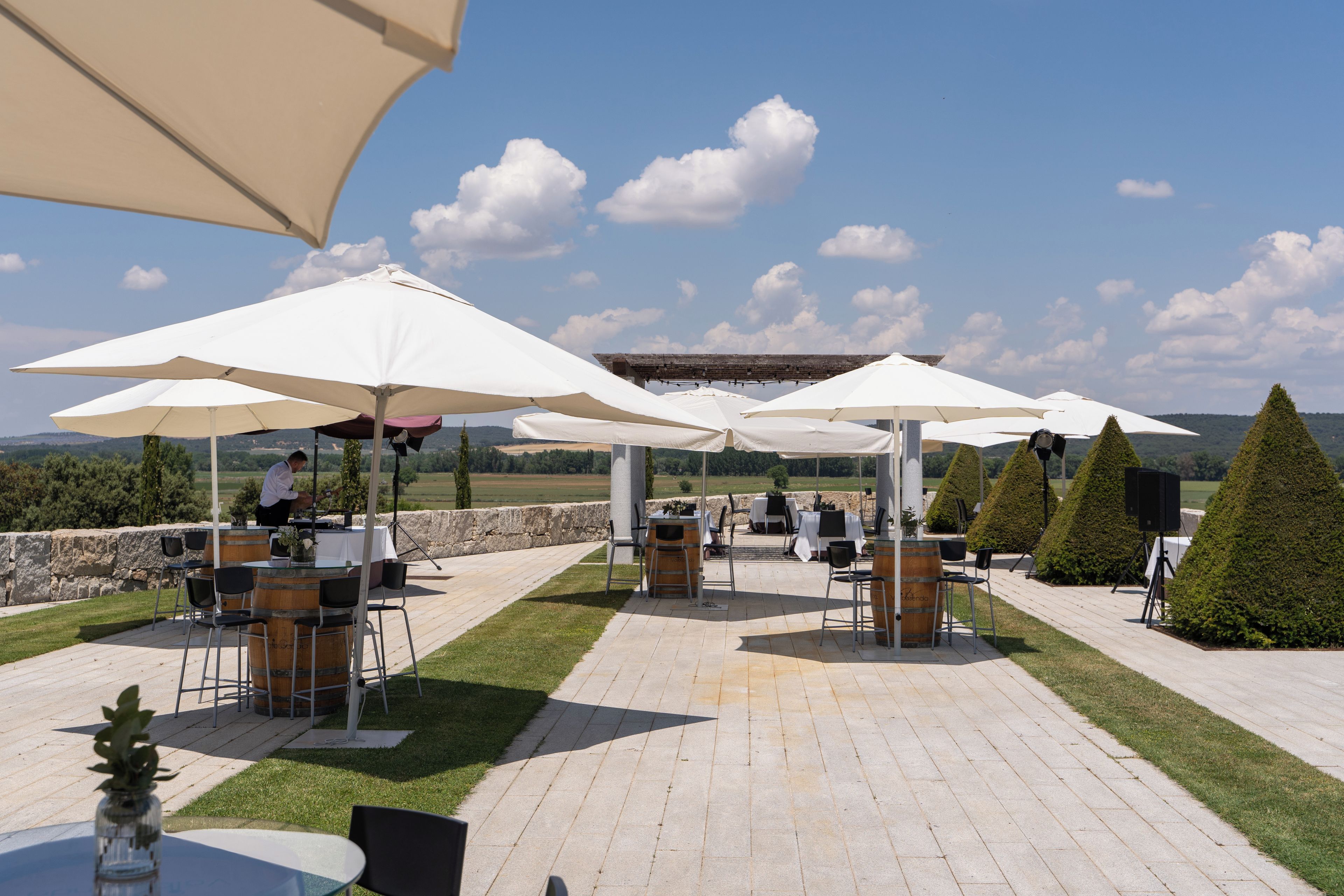An outdoor patio with white umbrellas, tables, and chairs, set against a backdrop of a stone wall and a clear sky.