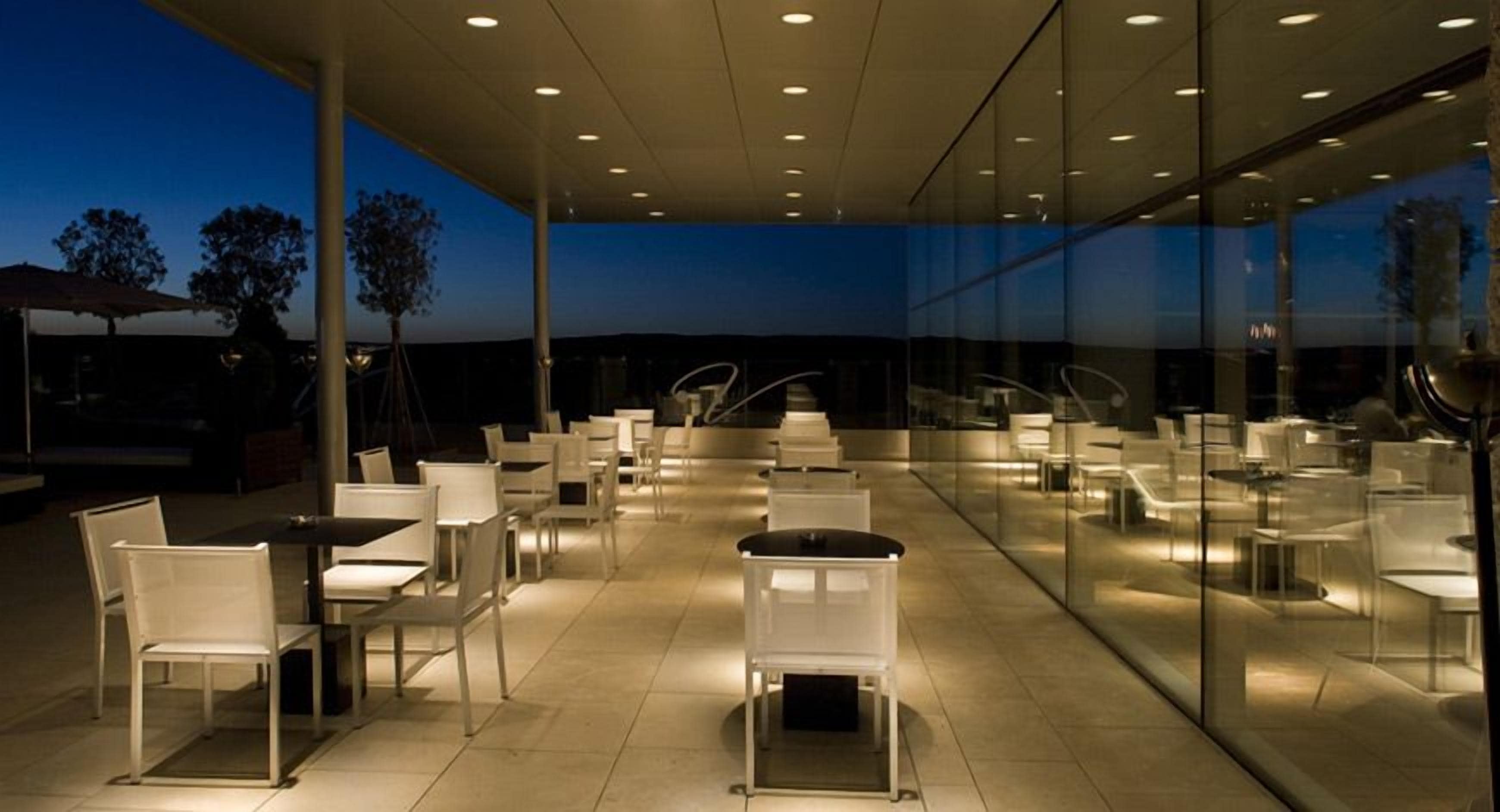 Un área de comedor al aire libre con sillas y mesas modernas, grandes ventanales de vidrio y vistas al cielo.