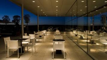 Un área de comedor al aire libre con sillas y mesas modernas, grandes ventanales de vidrio y vistas al cielo.