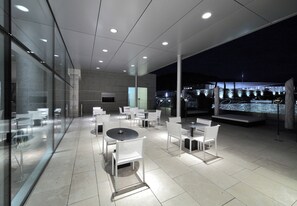 Zona d'exterior amb menjador modern, parets de vidre, mobiliari blanc i vistes a la ciutat de nit.