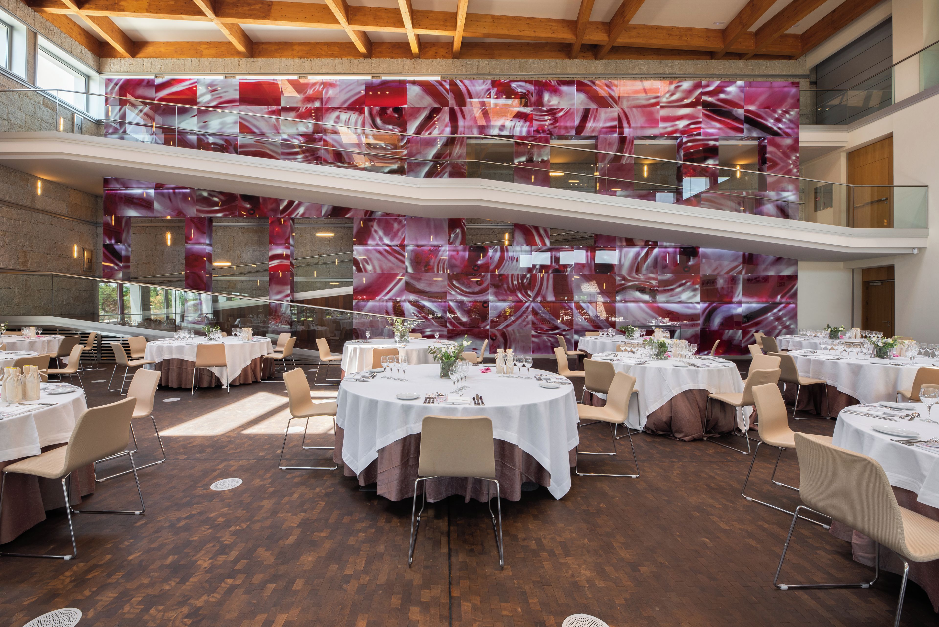 Un comedor de varios niveles con mesas redondas dispuestas para una comida, con un interior moderno, techo de madera y un gran mural abstracto en la pared superior.