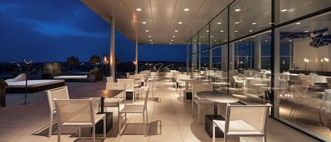 An outdoor dining area with modern furniture and glass walls, providing views of the cityscape at dusk.