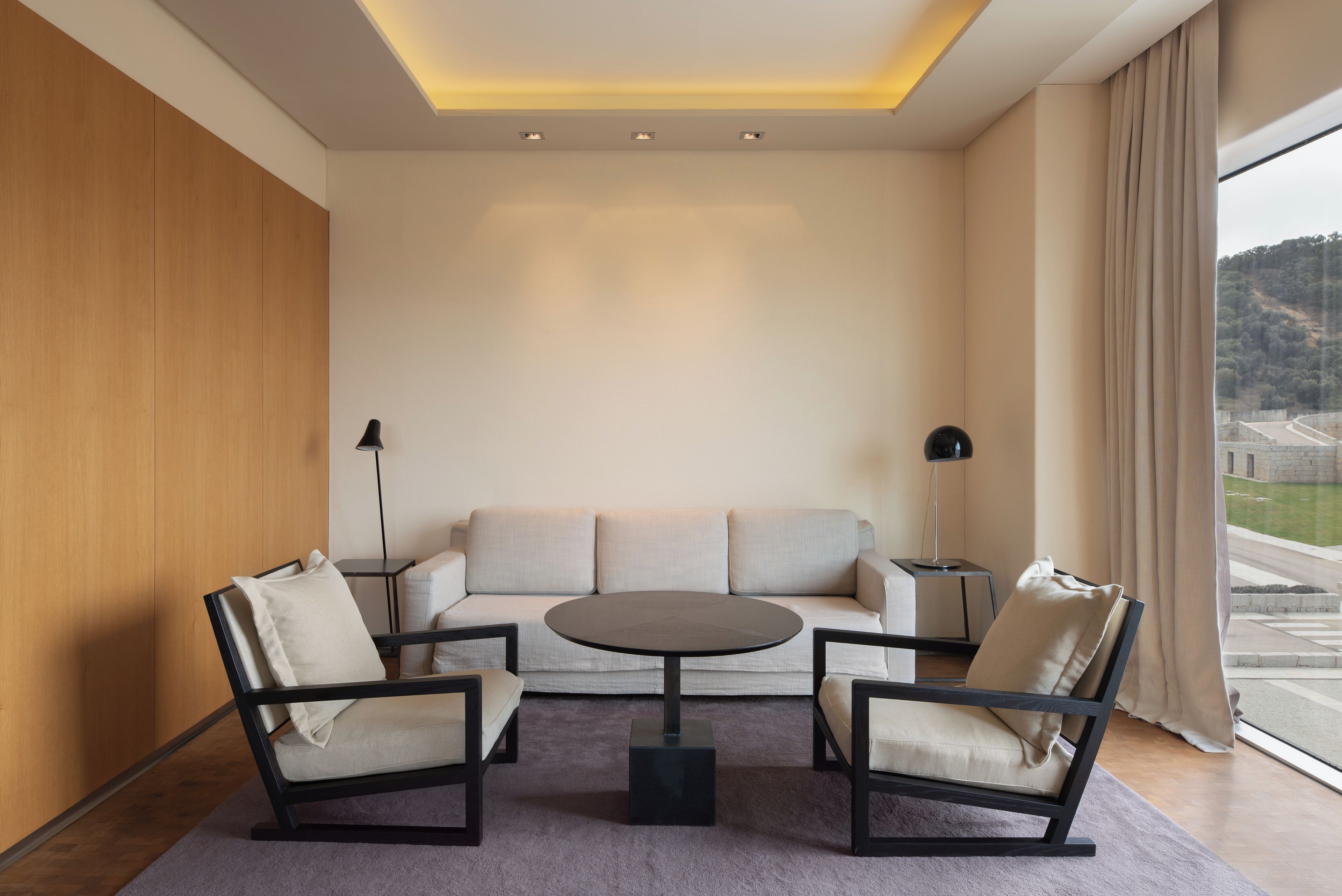 A modern living room with a sofa, two armchairs, a coffee table, and a view of a hilly landscape through large windows.