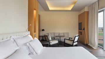 Habitación de hotel con una cama grande, un sofá, una mesita pequeña y sillas. Hay un televisor en la pared y una ventana con cortinas.