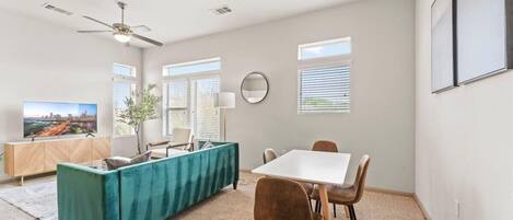 Una sala de estar moderna con un sofá color turquesa, una mesa de comedor con sillas, un ventilador de techo y ventanas grandes con persianas.