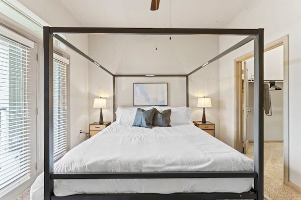 A four-poster bed with white bedding and two pillows, flanked by nightstands with lamps, in a room with a large window and a framed picture on the wall.