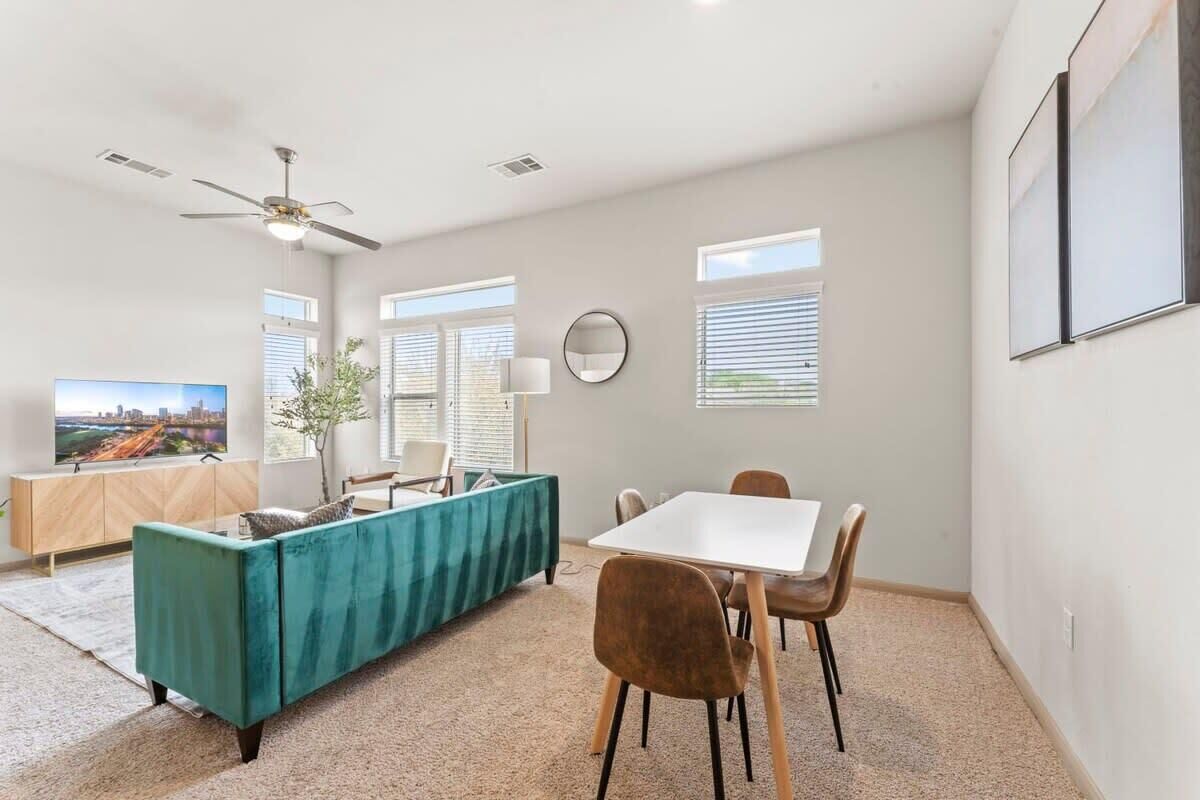 A modern living room with a teal sofa, a dining table with chairs, a ceiling fan, and large windows with blinds.