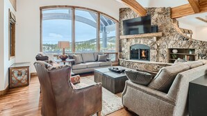 A spacious living room with a stone fireplace, leather furniture, and a large window offering a mountain view. - Free SkyCard Act-Lux Ski & Golf Villa, Hot Tub, Home Theater, Billiards, Wet Bar (Breckenridge)