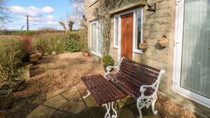 A stone house with a wooden bench in the front yard, a door with a knocker, and a window with a hanging pot. - The Old Croft (Buxton)