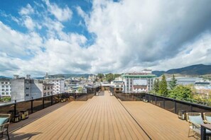 Eine Holzterrasse mit Tischen und Stühlen, mit Blick auf eine Stadtlandschaft aus Gebäuden und Bergen.