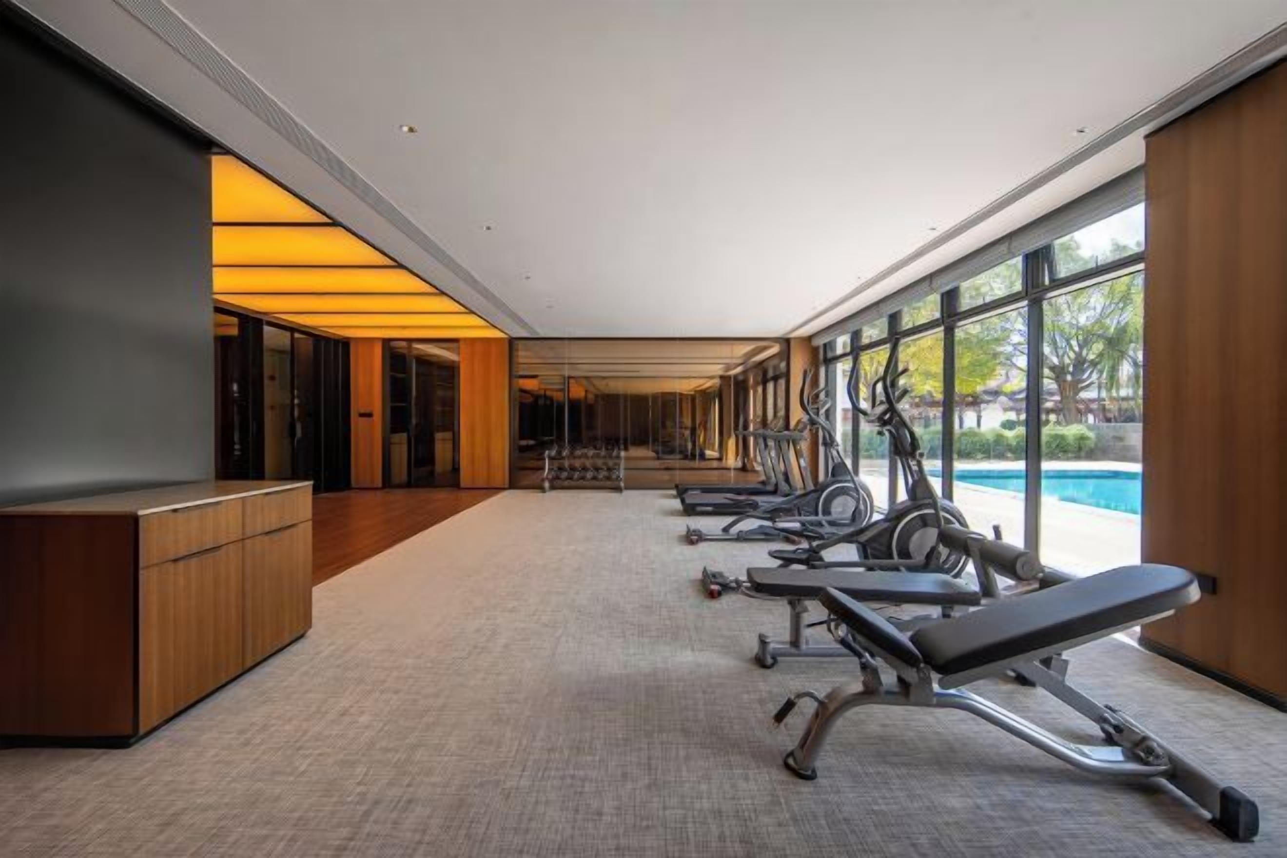 A gym with exercise equipment and a reception desk, large windows, and a view of a swimming pool.