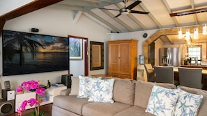 A living room with a beige sofa, a large painting of a beach, a wooden cabinet, and a kitchen area visible through an archway. - ST.JOHN, VIRGIN ISLANDS!  2 BEDROOM, 1.5 BATH, BEAUTIFUL TROPICAL GARDENS! (St. John)