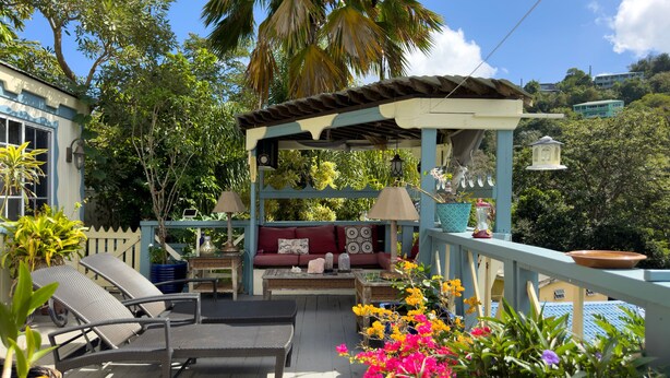 A covered outdoor seating area with wicker chairs and a cushioned bench, surrounded by lush greenery and a palm tree. - ST.JOHN, VIRGIN ISLANDS!  2 BEDROOM, 1.5 BATH, BEAUTIFUL TROPICAL GARDENS! (St. John)
