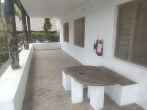 A covered outdoor area with a stone floor, white walls, a fire extinguisher mounted on the wall, and a bench.