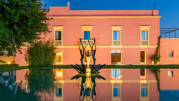 A pink building with a reflection in the water, a statue in the center, and a tree to the left. - CASIN ANAND (Novoli)