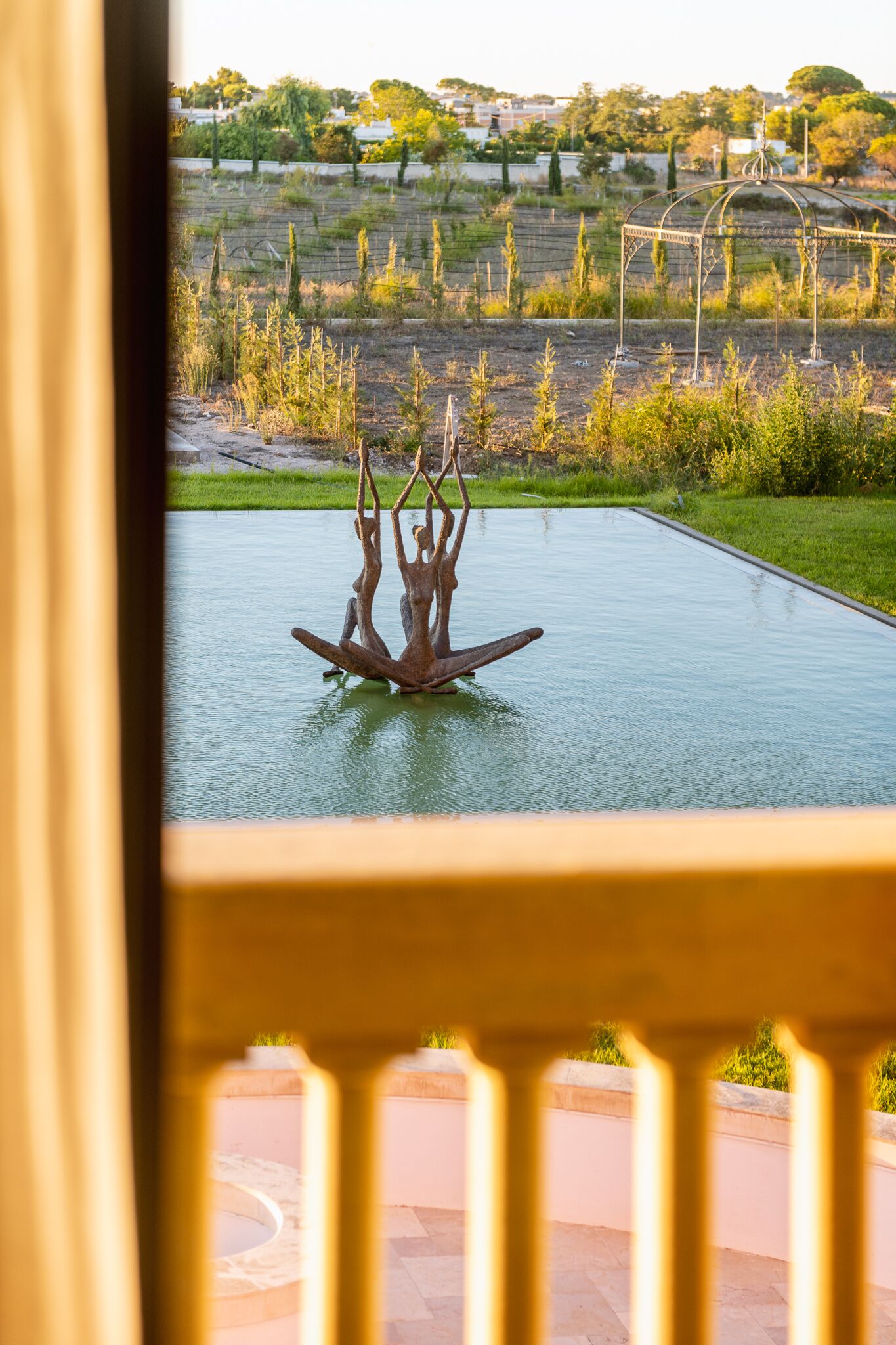 A view of a swimming pool with a sculpture in the center, surrounded by a landscaped garden and a vineyard in the background.