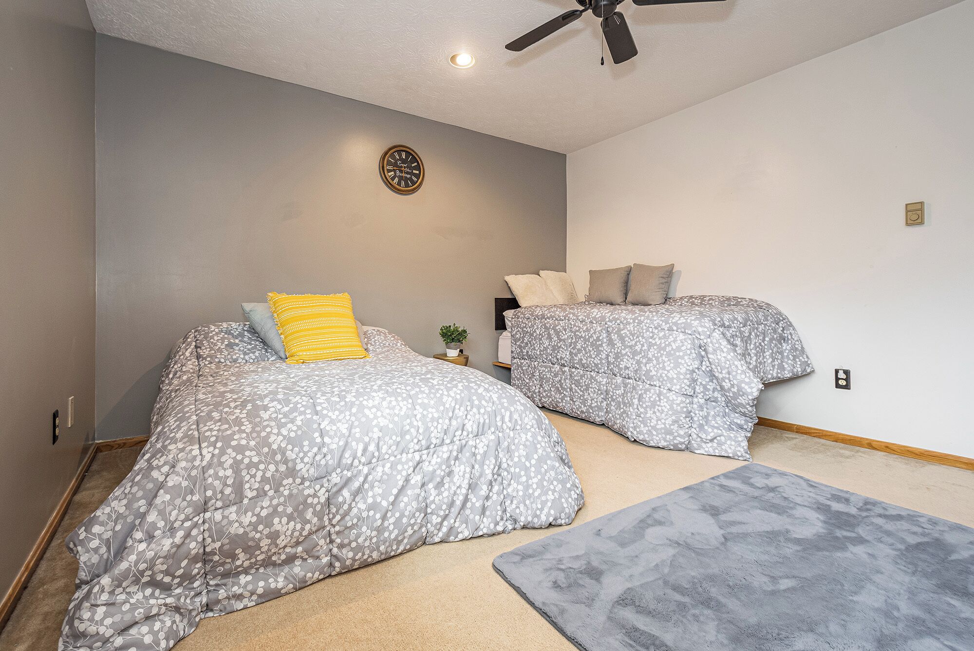 A bedroom with two beds, a ceiling fan, a wall clock, and a small plant.