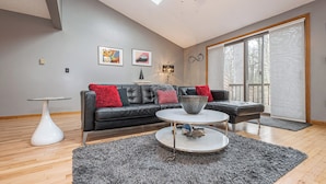 A modern living room with a black leather sofa, a round coffee table, and a grey shag rug. There is a large window with blinds, a floor lamp, and artwork on the wall. - Laurel Brook 39 by Taylor Made Deep Creek (Oakland)