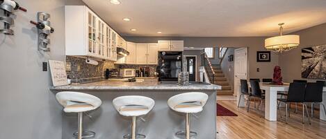Una cocina moderna con una isla central, taburetes de bar, un área de comedor con candelabro y una escalera que sube a otro nivel.