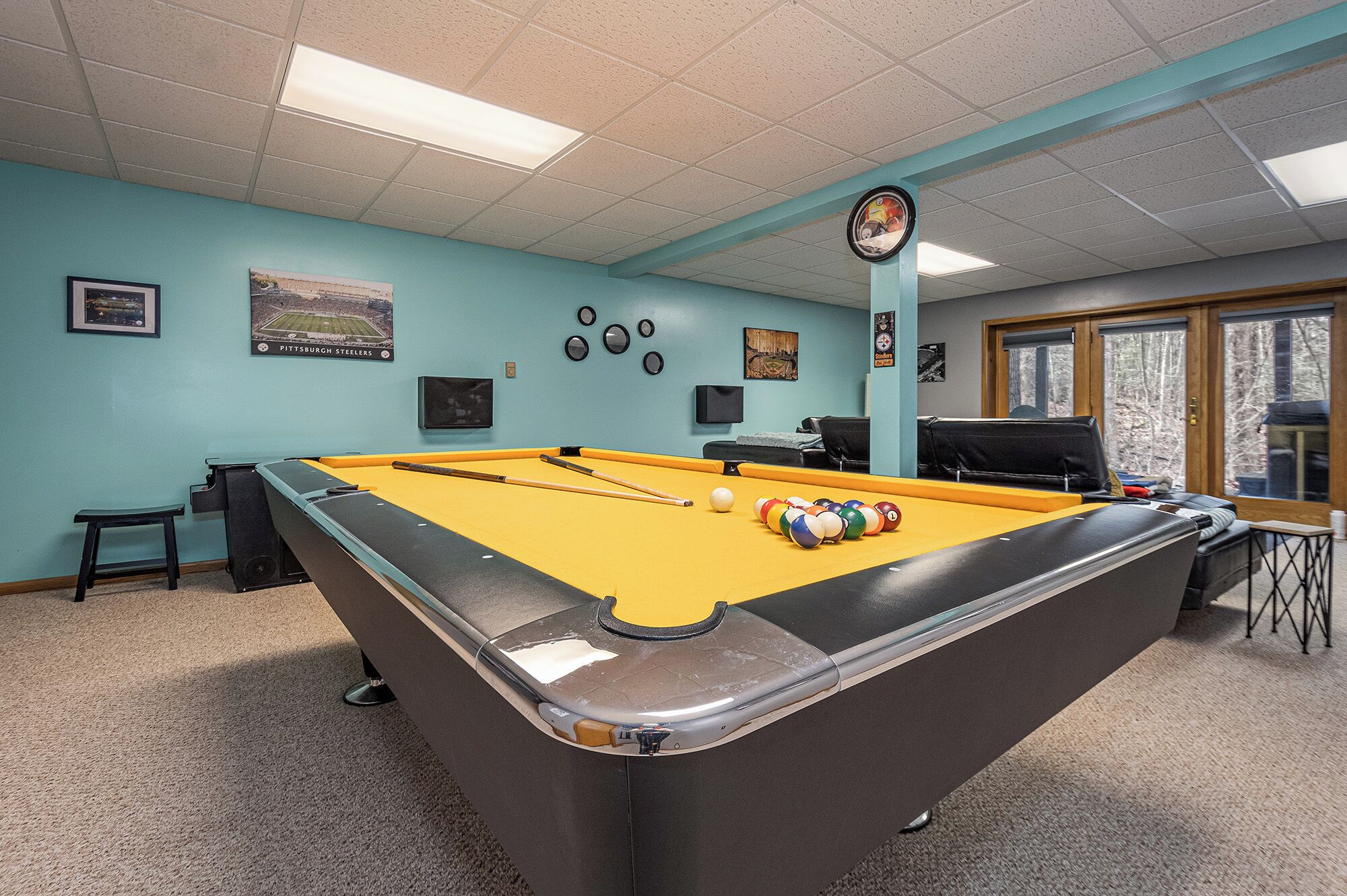 A pool table with a yellow felt surface, surrounded by teal walls, and a clock on the wall.