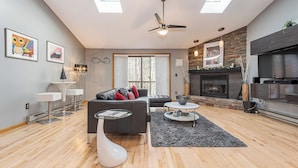 A modern living room with a fireplace, a ceiling fan, and a large window. - Laurel Brook 39 by Taylor Made Deep Creek (Oakland)