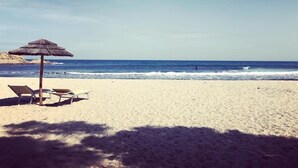 A beach with a thatched umbrella and two lounge chairs.