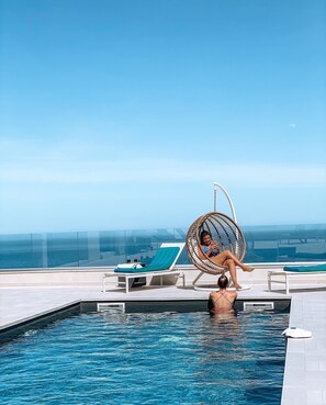 A pool area with a hanging chair and lounge chairs, overlooking a body of water.