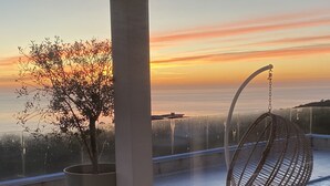 A wooden table and bench on a balcony with a hanging chair, overlooking a sunset with a tree and a clear sky.