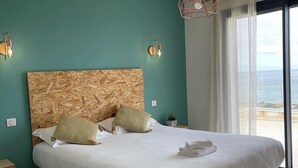 A bedroom with a large bed, two bedside tables, a headboard with a wood pattern, a hanging light fixture, and a view of the sea through a window.
