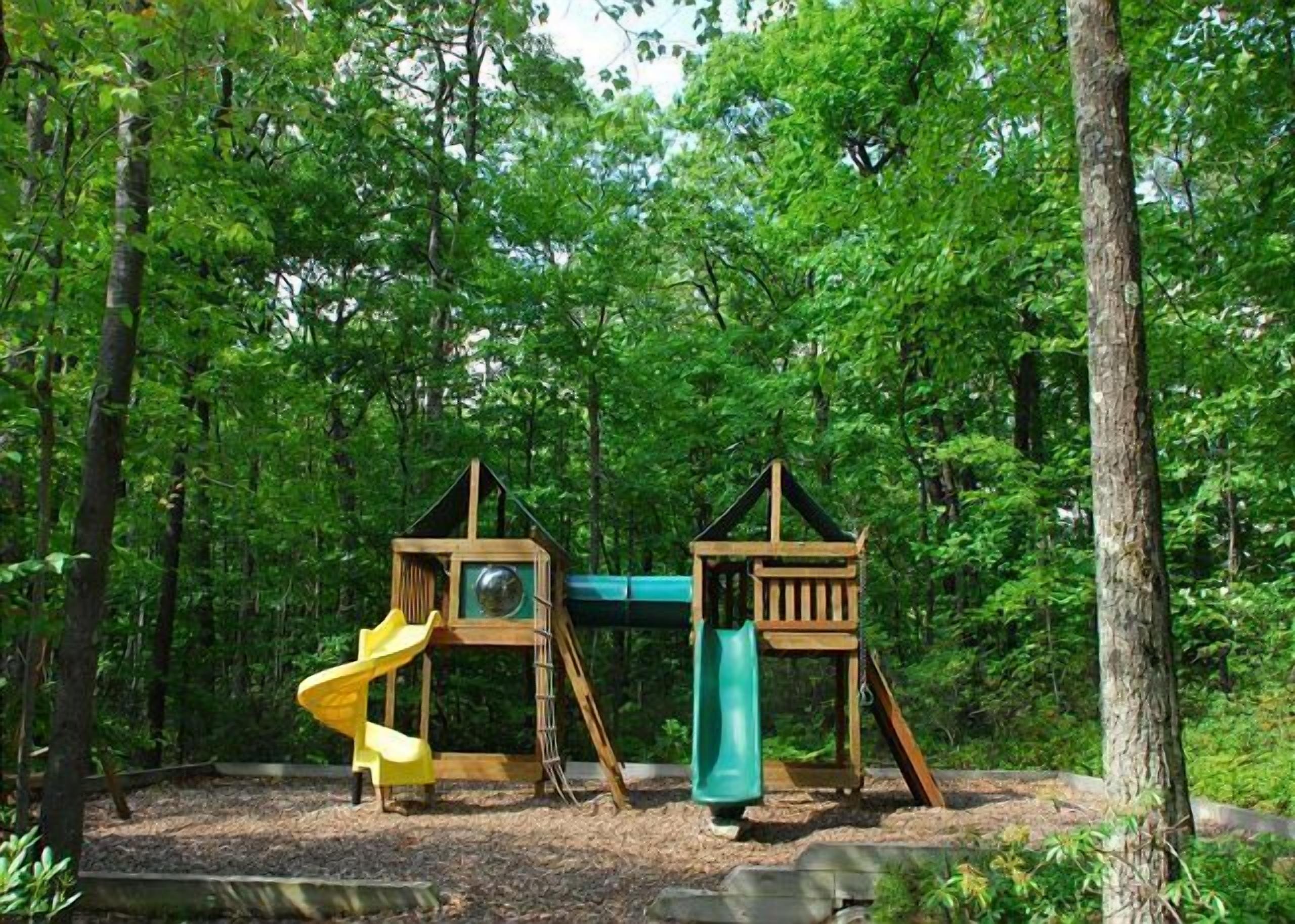 A wooden playground with two slides and a climbing structure in a forested area.